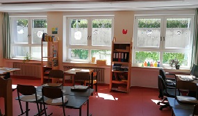 Ein Raum wird zur Fensterseite gezeigt. Dabei sieht man Bänke und Stühle. Rechts einen Computer.